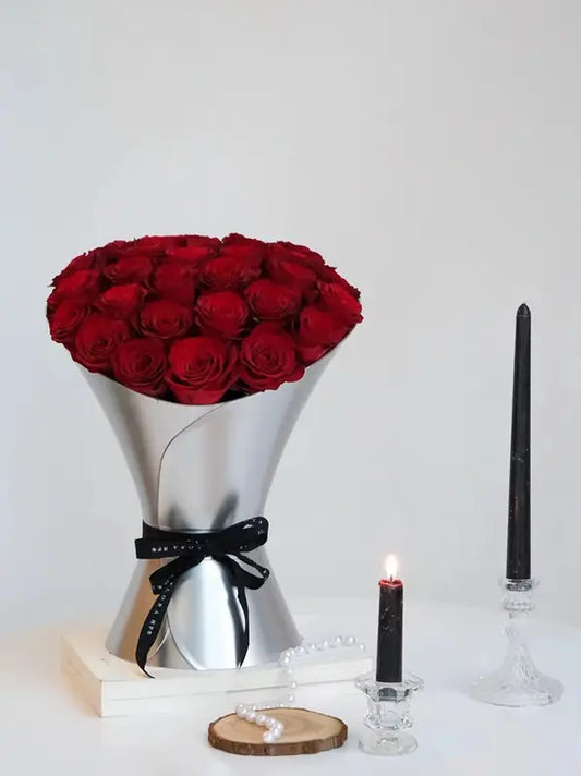 A cone-shaped hand held flower vase made from cardboard, presented with red roses inside. The vase appears to be silver in color, with a black ribbon decoration, placed on a wooden surface with two black candles in the background.