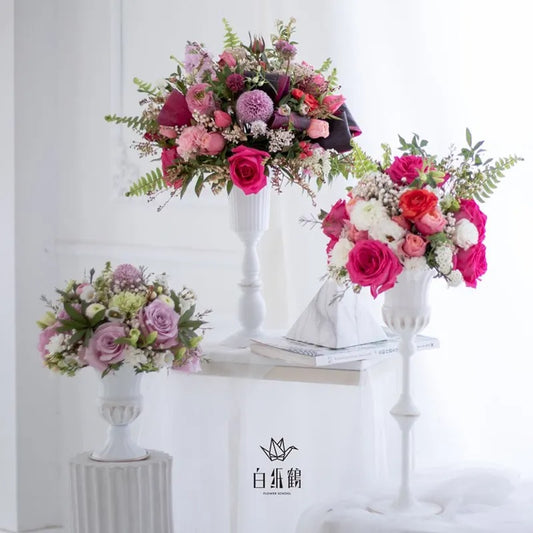 A set of vintage iron flower vases with wedding centerpieces arranged in them, displayed on a white surface. The vases are in gold, bronze, and white colors, accompanied by assorted flowers in pink, purple, and white.