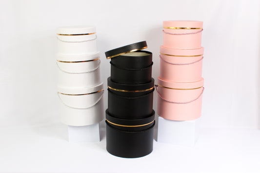 Stacked cylindrical gift boxes with gold rims in black, pink, and white colors.